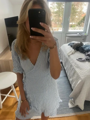 Blåvit omlottklänning med volang - Säljer en superfin kort omlottklänning med blå och vita ränder. Klänningen har v-ringning, knyt i midjan och volangdetaljer längs nederkanten. Perfekt för sommarens alla tillfällen och har en lätt och luftig känsla.