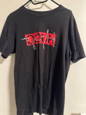 Svart HUGO t-shirt med tryck - Snygg svart t-shirt från HUGO med ett stort, grafiskt tryck i rött och silver på bröstet. Klassisk rund hals och korta ärmar. Perfekt för dig som gillar streetwear och vill ha en statement tee i garderoben.