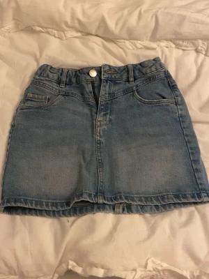 Blå jeanskjol med fickor - Säljer en klassisk blå jeanskjol i kort modell med fem fickor och bälteshällor. Kjolen har knapp och dragkedja framtill och är gjord i ett mjukt denimtyg. 