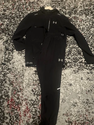 Svart träningsset från Under Armour vanish 2.0 - Säljer ett svart träningsset från Under Armour med jacka och byxor. Setet har vita loggor och detaljer, dragkedja på jackan och elastisk midja på byxorna. Perfekt för löpning eller gym. Materialet känns lätt och andas bra.