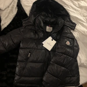  Moncler Jacka - Säljer en svart pufferjacka från Moncler med avtagbar huva och klassisk logga på ärmen. Jackan har dragkedja framtill, två sidofickor med dragkedja och quiltad design. Insidan har ett unikt serietidningsmärke. Perfekt för kalla dagar och riktigt snygg streetstil. Storlek 1 alltså S i vanlig storlek.