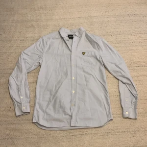 Ljusblå skjorta från Lyle & Scott - Snygg ljusblå skjorta från Lyle & Scott med klassisk krage och knappar framtill. På bröstet finns en ficka med den ikoniska gula örnloggan. Skjortan är långärmad och tillverkad i mjuk bomull, perfekt för en clean och stilren look.