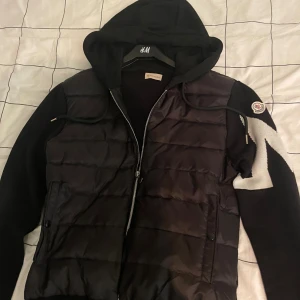 Svart Moncler pufferjacka med huva - Svart pufferjacka från Moncler med huva och dragkedja. Jackan har stickade ärmar med vit zigzag-detalj och Moncler-logga på ena ärmen. Kroppen är quiltad och fylld med dun för extra värme. Snygg och sportig look, perfekt för kyliga dagar. Kvitto finns att se