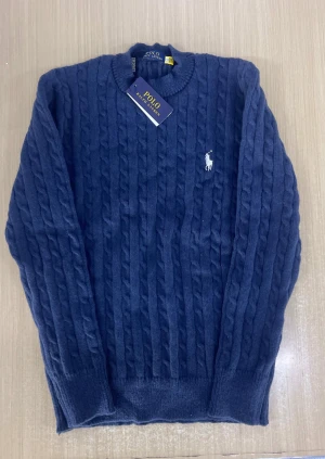 Marinblå stickad tröja Polo Ralph Lauren - Snygg marinblå stickad tröja från Polo Ralph Lauren med klassiskt kabelstickat mönster och rund hals. Tröjan har långärmade ärmar och den ikoniska broderade loggan i vitt på bröstet. Perfekt för dig som gillar stilren och tidlös design.