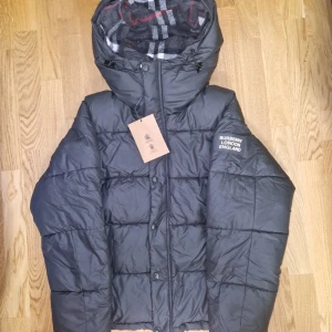 Burberry jacka - Snygg svart dunjacka från Burberry med klassisk logga på ärmen och rutigt foder i huvan. Jackan har dragkedja, knappar och justerbar huva. Perfekt för kalla dagar och riktigt stilren look.