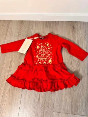 Röd julklänning med volanger Choupette - Supersöt röd kortklänning från Choupette med guldiga stjärnor och texten 'Be Merry and Bright' framtill. Klänningen har långa ärmar, rund hals och två lager volanger på kjolen. Perfekt för julens alla tillfällen och tillverkad i mjuk bomull.