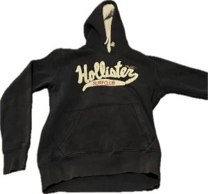 Svart hoodie från Hollister Surfclub - Svart hoodie från Hollister Surfclub med stor broderad logga framtill. Tröjan har huva med vit snörning och mjukt foder, samt en klassisk känguruficka. Perfekt för dig som gillar en avslappnad och sportig stil.
