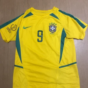 Brasilien Ronaldo VM 2002 tröja Nike S - Gul Brasilien fotbollströja från VM 2002 med gröna detaljer, nummer 9 och Ronaldo på ryggen. Nike-logga på bröstet och officiellt FIFA World Cup Korea Japan-tryck på ärmen. Ventilerande meshpaneler och klassisk rund hals. Perfekt för fans av legenden Ronaldo.