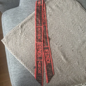 Röd och brun Fendi halsduk i siden - Supersnygg smal halsduk från Fendi i rött och brunt med tydliga loggor och texten 'ROMA AMOR'. Gjord i lyxigt siden, perfekt att knyta runt halsen eller väskan för en cool statement-look. Designen har grafiskt mönster och kontrasterande färger.