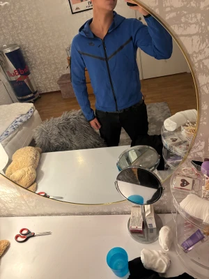 Blå Nike tech hoodie från JD med dragkedja - Blå Nike tech hoodie från JD med svarta detaljer och hel dragkedja framtill. Tröjan har huva, långärmade ärmar och en sportig look med svarta linjer över bröstet och på ena ärmen. Perfekt för dig som gillar streetwear och sportig stil.