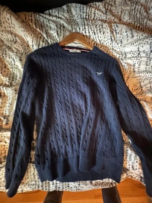 En blå gant över tröja - Snygg marinblå kabelstickad tröja från Hampton Republic med rund hals och diskret logga på bröstet. Tröjan har långärmad passform och ribbade muddar vid ärmslut och nederkant. Perfekt för dig som gillar klassisk stil och vill ha något som funkar året runt.