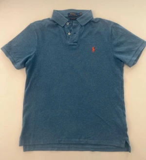 Blå pikétröja från Polo Ralph Lauren - Snygg blå pikétröja från Polo Ralph Lauren i bomull, med klassisk krage och två vita knappar framtill. Custom slim fit för en modern siluett. Orange broderad logga på bröstet ger en stilren touch. Perfekt för dig som gillar enkel och clean stil. Skicket är mycket bra 
