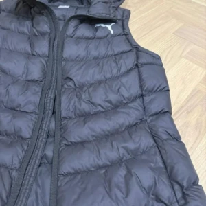 Svart dunväst med huva från Puma - Svart dunväst från Puma med huva och dragkedja framtill. Västen har ett quiltat mönster och Puma-logga i vitt på bröstet och ryggen. Perfekt för kyliga dagar när du vill ha en sportig och clean look.