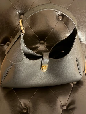 Svart axelväska med gulddetalj - Stilren svart axelväska i skinn med snygg guldfärgad låsdetalj framtill. Väskan har en mjukt rundad form och bred axelrem med guldfärgat spänne. Perfekt storlek för att få plats med allt du behöver och ger en clean vibe till din outfit.