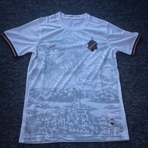 Vit AIK fotbollströja Nike  - AIK fotbollströja i vitt med detaljer i guld och svart, tillverkad i ventilerande polyester. Tryckt stadsmotiv över hela framsidan, AIK-emblem på bröstet och Nike-logga i guld. Kortärmad modell med svarta och guldiga ränder vid ärmslut. Texten STOCKHOLMENSIS i nacken.