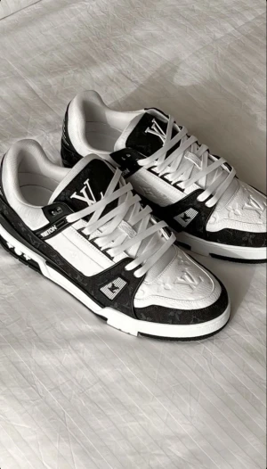 Louis Vuitton svartvita sneakers - Snygga Louis Vuitton sneakers i svart och vitt med klassiskt monogrammönster. Skorna har rund tå, platt sula och är tillverkade i skinn och textil. De har vita snören och coola LV-detaljer på sidan och tungan. Perfekt för dig som gillar exklusiv streetstyle.