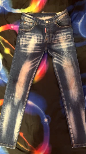 Dsquared2 blå skinny jeans med tryck - Snygga blå jeans från Dsquared2 med blekta detaljer och stort DSQUARED2-tryck både fram och bak. Skinny fit med klassisk femficksdesign, knappgylf och coolt sliten look. Perfekt för dig som gillar streetstyle och vill sticka ut.