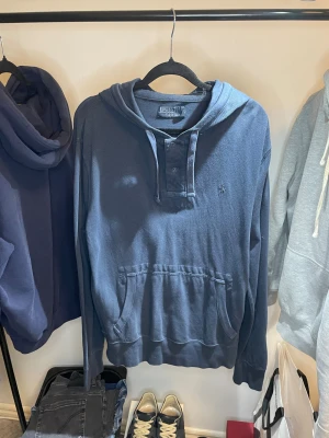 Blå hoodie från Polo Ralph Lauren - Snygg marinblå hoodie från Polo Ralph Lauren med huva, knappar vid halsen och klassisk logga broderad på bröstet. Tröjan har en stor magficka och är tillverkad i mjuk bomull. Perfekt för chill dagar och enkel att matcha med jeans eller joggers.