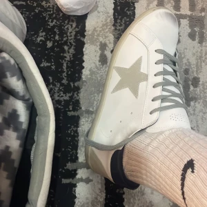 GGDB Superstar vita sneakers stl 43 - Säljer ett par GGDB Superstar sneakers i vitt skinn med grå snörning och beige stjärndetalj på sidan. Skorna har en mörkblå häl och platt sula. Klassisk och cool design med kontrastdetaljer, perfekt för dig som gillar streetstyle.