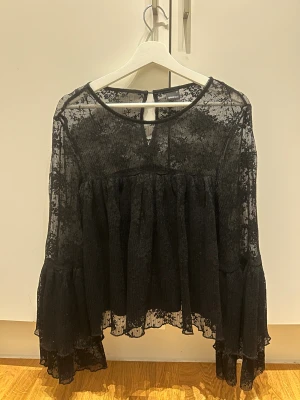 Svart spetsblus med volang från Gina Tricot - Supersnygg svart blus i transparent spets från Gina Tricot. Blusen har långa ärmar med breda volanger och ett blommigt spetsmönster. Rund halsringning med knapp i nacken och flowy passform. Perfekt för dig som gillar statement-plagg!