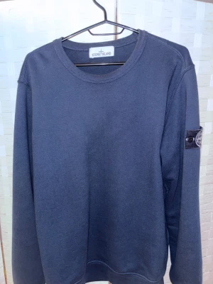 Svart sweatshirt från Stone Island - Svart sweatshirt från Stone Island med klassisk rund hals och lång ärm. Tröjan har den ikoniska Stone Island-loggan på vänster ärm, vilket ger en clean och stilren look. Materialet är mjukt och känns skönt mot huden.