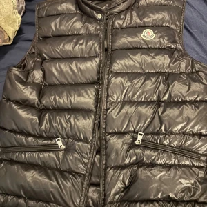 Svart dunväst från Moncler - Snygg svart dunväst från Moncler med quiltad design och hög krage. Västen har två praktiska fickor med dragkedja och klassisk Moncler-logga på bröstet. Perfekt för lager-på-lager och ger en clean, sportig look. Stängs med dragkedja framtill. Går att prata om priset 