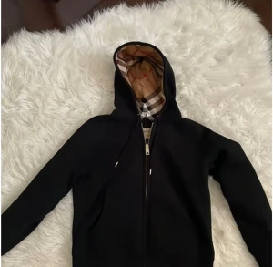 Svart Burberry hoodie med dragkedja - Svart hoodie från Burberry med klassisk rutig huva i beige, svart och röd. Tröjan har hel dragkedja framtill, snörning vid huvan och långa ärmar. Märket syns tydligt på insidan och detaljerna är stilrena. Perfekt för dig som vill ha en snygg streetwear-look. Skulle säga att den passar M och runt den storleken!