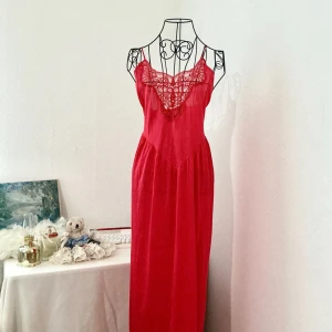 Röd lång nattklänning med spetsdetaljer - Snygg röd långklänning i nylon med tunna axelband och vacker spets upptill. Materialet är tunn så den är något genomskinlig. Storlek:L, passar storlek 40-42. Första bild visar hur det ser ut ungefär på en kropp strl. 40-42. Andra bilder visar hur den ser ut på dockan som har storlek 34.