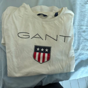 Vit GANT t-shirt med tryck - Vit t-shirt från GANT med stort tryck på bröstet och ett coolt emblem med stjärnor och röda ränder. Klassisk rund hals och korta ärmar. Mjuk och skön bomull som passar perfekt till jeans eller shorts.