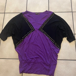 Lila och svart topp med nitar - Snygg lila och svart topp med korta ärmar och v-ringning. Toppen har dekorativa nitar längs de svarta partierna som ger en cool look. Perfekt för en trendig stil.