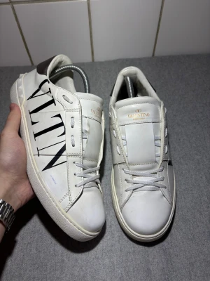 Valentino Garavani skor - Snygga vita Valentino Garavani sneakers i skinn med svart VLTN-logga på sidan och coola nitar runt hälen. Skorna har snörning, rund tå och en silvrig detalj på baksidan. Perfekta för dig som vill sticka ut med exklusiva och trendiga sneakers.