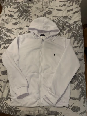 Vit Polo Ralph lauren kofta  - Vit hoodie från Polo Ralph Lauren med dragkedja och klassisk broderad logga på bröstet. Tröjan har huva, två fickor framtill och är tillverkad i mjuk bomull. Perfekt för dig som gillar clean och stilren look.