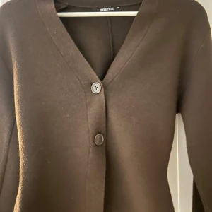 Brun kofta från Gina Tricot - Mysig brun kofta från Gina Tricot med v-ringad hals och stora knappar framtill. Koftan har långa ärmar och är tillverkad i ett mjukt material som känns skönt mot huden. Perfekt att slänga på sig när det är lite kyligt ute.