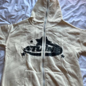 Beige hoodie från 4Tune med tryck - Säljer en beige hoodie från 4Tune med svart grafiskt tryck och stjärnor både fram och bak. Tröjan har huva, hel dragkedja och stora fickor framtill. Perfekt för dig som gillar streetwear och snygga detaljer.