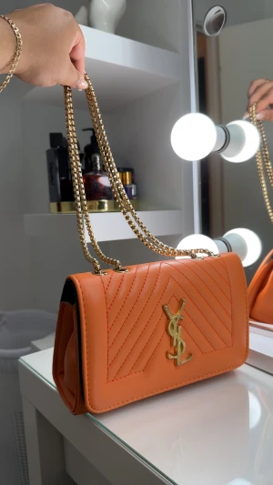 Orange YSL axelväska med kedja - Snygg orange axelväska från YSL med guldfärgad kedja och ikonisk YSL-logga framtill. Väskan är quiltad med ett stilrent mönster och har en rektangulär form. Perfekt för dig som vill sticka ut med en lyxig accessoar.