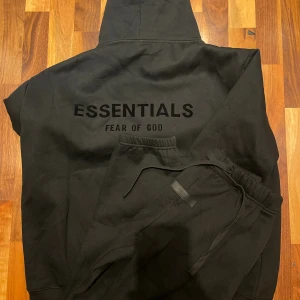 Svart Essentials Fear of God tracksuit - Svart tracksuit från Essentials Fear of God med stor logga på ryggen och mindre logga på bröstet. Klassisk känguruficka, huva och ribbade muddar. Oversized passform och tillverkad i mjuk bomull, perfekt för en chill och stilren look. Byxlängd: 105cm midjebredd: 36cm
