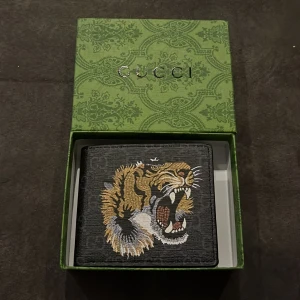 Svart Gucci plånbok med tigerbroderi - Snygg svart plånbok från Gucci med broderad tiger på framsidan och diskret GG-mönster. Plånboken har flera kortfack och fönster för ID. Tillverkad i exklusivt skinn med lyxig känsla och levereras i original grön Gucci-box.
