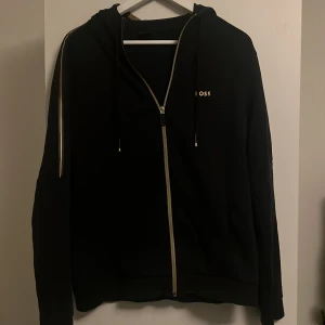 Svart hoodie från BOSS med dragkedja - Svart hoodie från BOSS med gulddetaljer och dragkedja framtill. Tröjan har huva med snörning och långa ärmar. Snygg guldfärgad rand längs axlarna och BOSS-logga på bröstet. Perfekt för chill eller streetwear.
