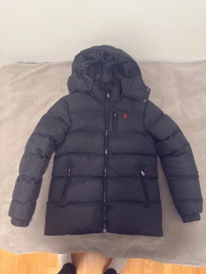 Ralph Lauren puffer svart - Svart pufferjacka från Polo Ralph Lauren med röd broderad logga på bröstet. Jackan har huva, tre dragkedjeförsedda fickor och elastiska muddar vid ärmslut. Materialet är syntet och jackan är quiltad för extra värme. Perfekt för kalla dagar.  PRIS KAN DISKUTERAS