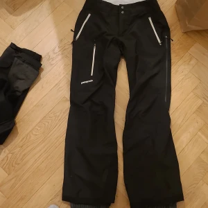 Svarta täckbyxor från Patagonia dam M - Svarta täckbyxor från Patagonia i damstorlek M. Byxorna har flera dragkedjeförsedda fickor framtill och är tillverkade i slitstark polyester. Perfekta för vintern och outdoor-aktiviteter. Snygg, enkel design med logga på benet och justerbara benslut.