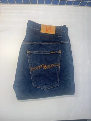 Nudie jeans - Snygga mörkblå jeans från Nudie Jeans med klassisk orange söm på bakfickorna och Nudie-logga på läderpatchen. Modellen är slim fit. Storlek och etikett lapp saknas. Jag bedömer storleken till 32/34 men utgå ifrån måtten ! JAG SKICKAR EJ FLER BILDER NÄR JAG BÄR JEANSEN! för storleksguide hänvisar jag till måtten och för bättre inblick i passformen rekommenderar jag att googla på modellnamnet. :)  47