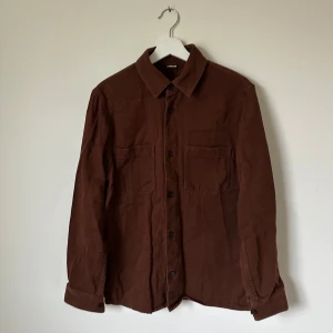 Brun overshirt från Arket i bomull - Brun overshirt från Arket i ekologisk bomull. Skjortan har klassisk krage, två stora bröstfickor och knappar framtill. Perfekt att slänga över en t-shirt för en chill look. Materialet är mjukt och lite tjockare, vilket gör den extra skön. 5J3