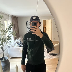 Grön hoodie från Hugo Boss med dragkedja - Snygg mörkgrön hoodie från Hugo Boss med svarta detaljer på axlarna och en vit logga på bröstet. Tröjan har huva, hel dragkedja och två fickor framtill. Perfekt för dig som gillar stilrena och sportiga plagg med premiumkänsla.