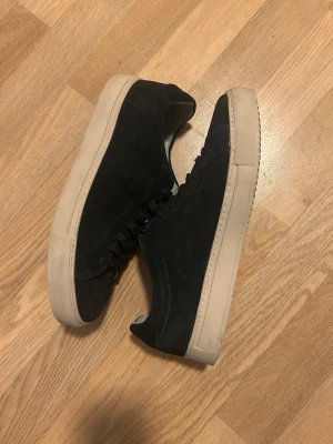 Svarta sneakers från Bugatti, strl 43 - Snygga svarta sneakers från Bugatti i mocka med beige platt sula. Klassisk och stilren design med rund tå och svarta snören. Perfekta för dig som gillar minimalistisk stil och vill ha ett par schyssta skor till vardags.