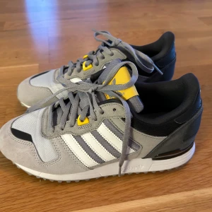  Nya Adidas sneakers i mocka i grått och vitt - Snygga Adidas sneakers i grått, vitt och svart med gula detaljer. Skorna har klassiska tre ränder på sidan, snörning och en mix av mocka och syntetmaterial. Perfekta för dig som gillar retrostil och bekväma skor med coola färgdetaljer.