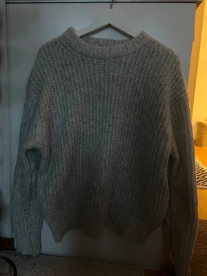 Grå stickad tröja med grov struktur - Mysig grå stickad tröja med grov ribbad struktur och rund hals. Tröjan har långärmade ärmar och en loose passform som ger en chill vibe. Perfekt för att hålla sig varm under kyliga dagar. Enkel att matcha med jeans eller leggings. (aningen oversize)
