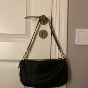 Svart axelväska från NLY Accessories (Nelly)  - Snygg svart axelväska från NLY Accessories med stilren, rundad form och guldfärgad dragkedja. Perfekt storlek. Mycket bra skick. Endast använd ett fåtal gånger. Köparen står för frakten!