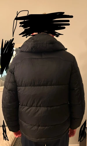 Svart pufferjacka från Polo Ralph Lauren - Svart pufferjacka från Polo Ralph Lauren med röd logga på bröstet. Jackan har huva, dragkedja framtill och två sidofickor med dragkedja. Den är quiltad och har en normal passform med långa ärmar. Perfekt för kalla dagar.