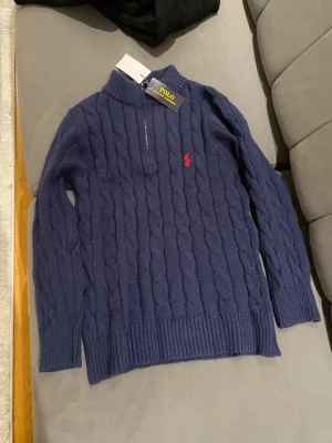 Marinblå stickad tröja Polo Ralph Lauren - Snygg marinblå stickad tröja från Polo Ralph Lauren med kabelstickat mönster och röd logga på bröstet. Tröjan har lång ärm, ribbade muddar och en half zip vid halsen för extra stilpoäng. Perfekt för dig som gillar klassisk och clean look.