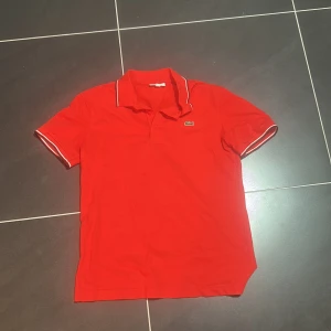 Orange pikétröja från Lacoste Sport - Orange pikétröja från Lacoste Sport med klassisk krage och vita detaljer på krage och ärmslut. Ikoniska krokodilloggan på bröstet. Tröjan är i bomull och har korta ärmar, perfekt för en sportig och stilren look. Använd 1 gång